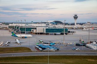 Flughafengelände mit einem Kontrollturm und parkenden Flugzeugen; im Vordergrund stehen Busse und Servicefahrzeuge, während ein Passagierflugzeug auf dem Rollfeld positioniert ist